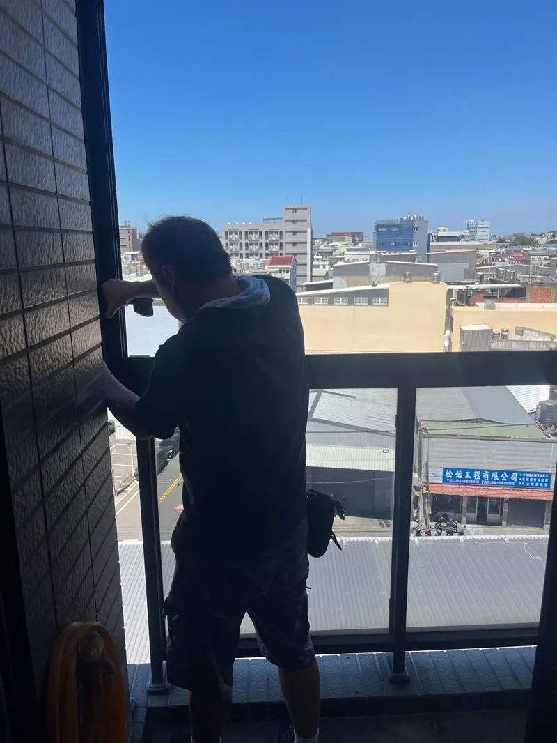 師傅站在陽台窗框旁施作鋁門窗細節，窗外是新豐一帶的街景與藍天